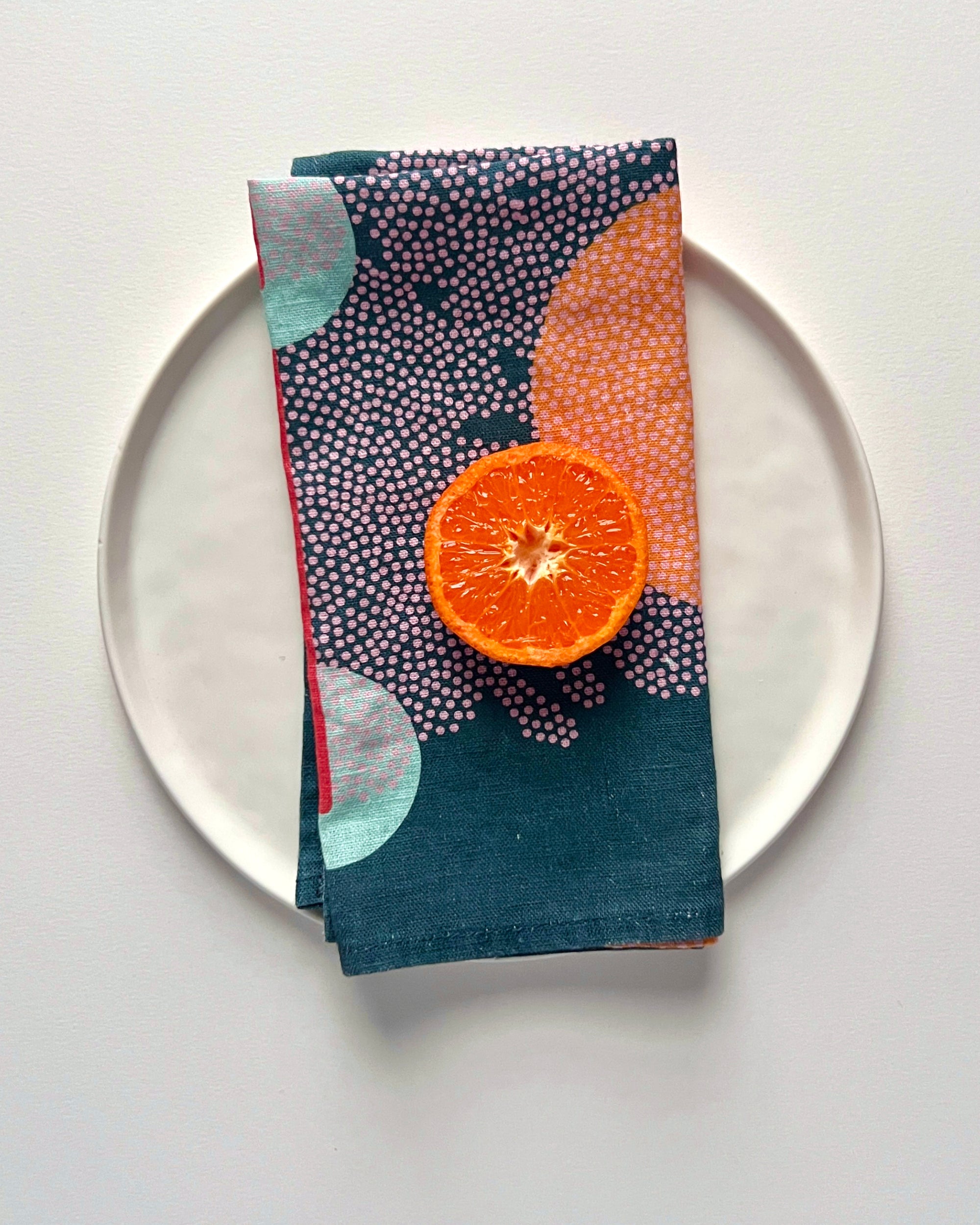 A blue linen napkin with an abstract pattern, displayed on a white plate alongside a halved orange. Designed by artist Leila Simon Hayes. 