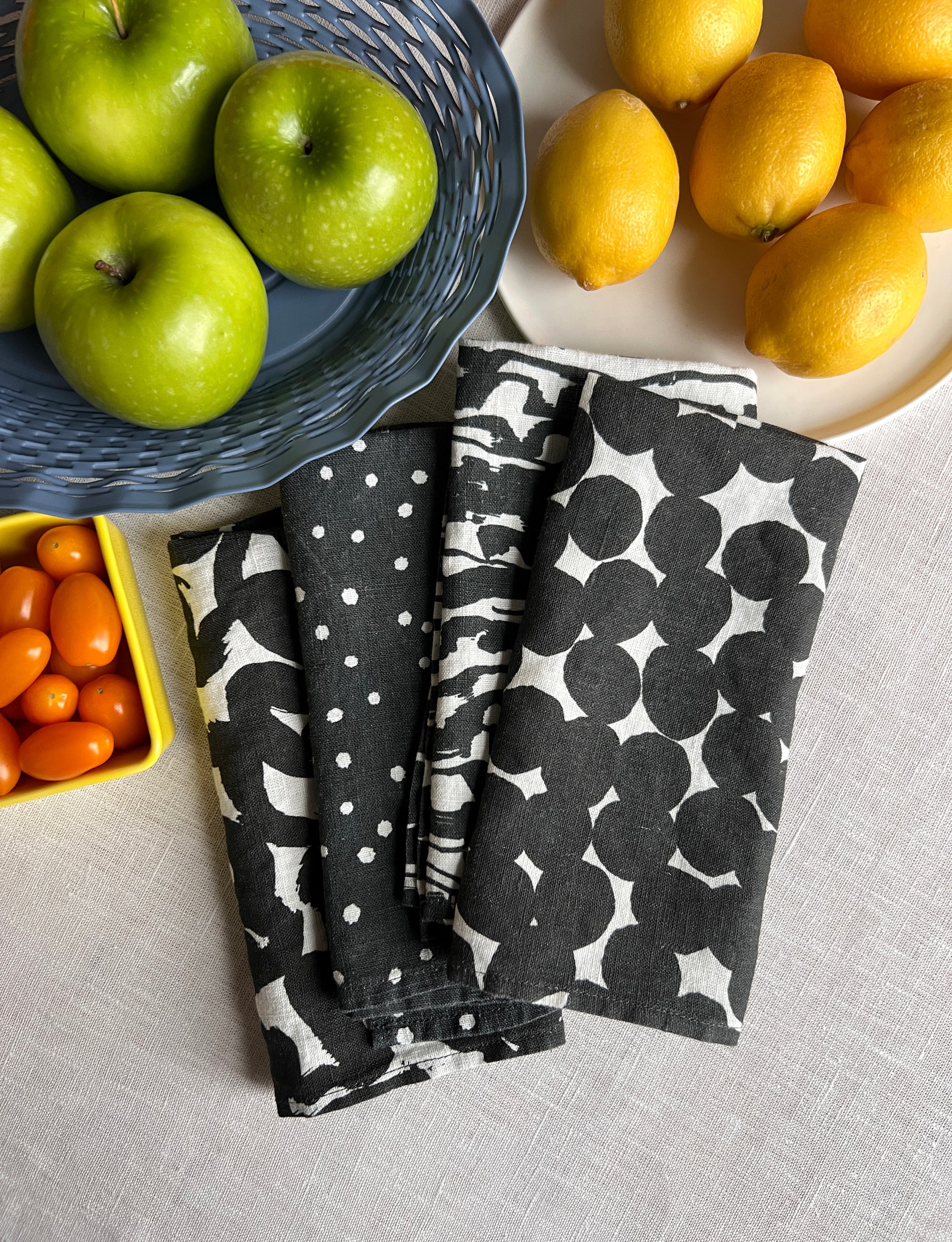 A set of four black and white linen napkins with a geometric pattern, displayed alongside a green bowl with two lemon halves. Designed by artist Leila Simon Hayes.