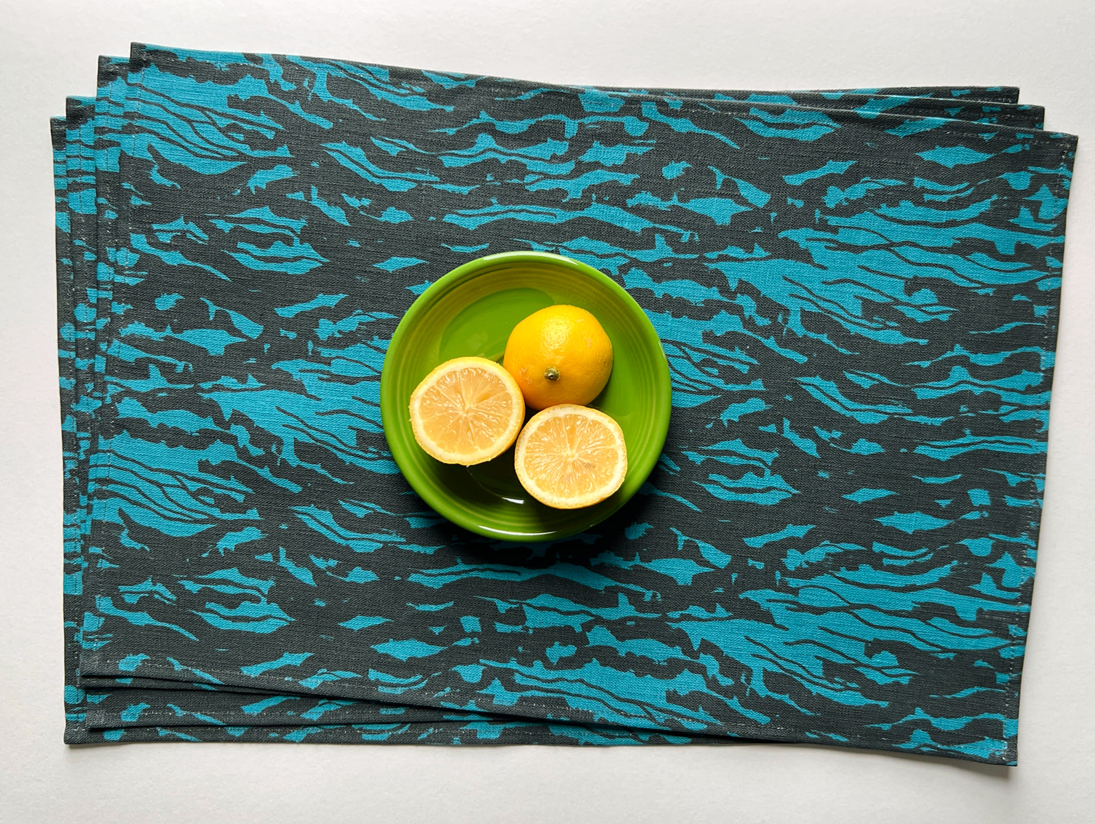 A blue linen placemat with a wave pattern, displayed with a green bowl and lemon halves on top. Designed by artist Leila Simon Hayes