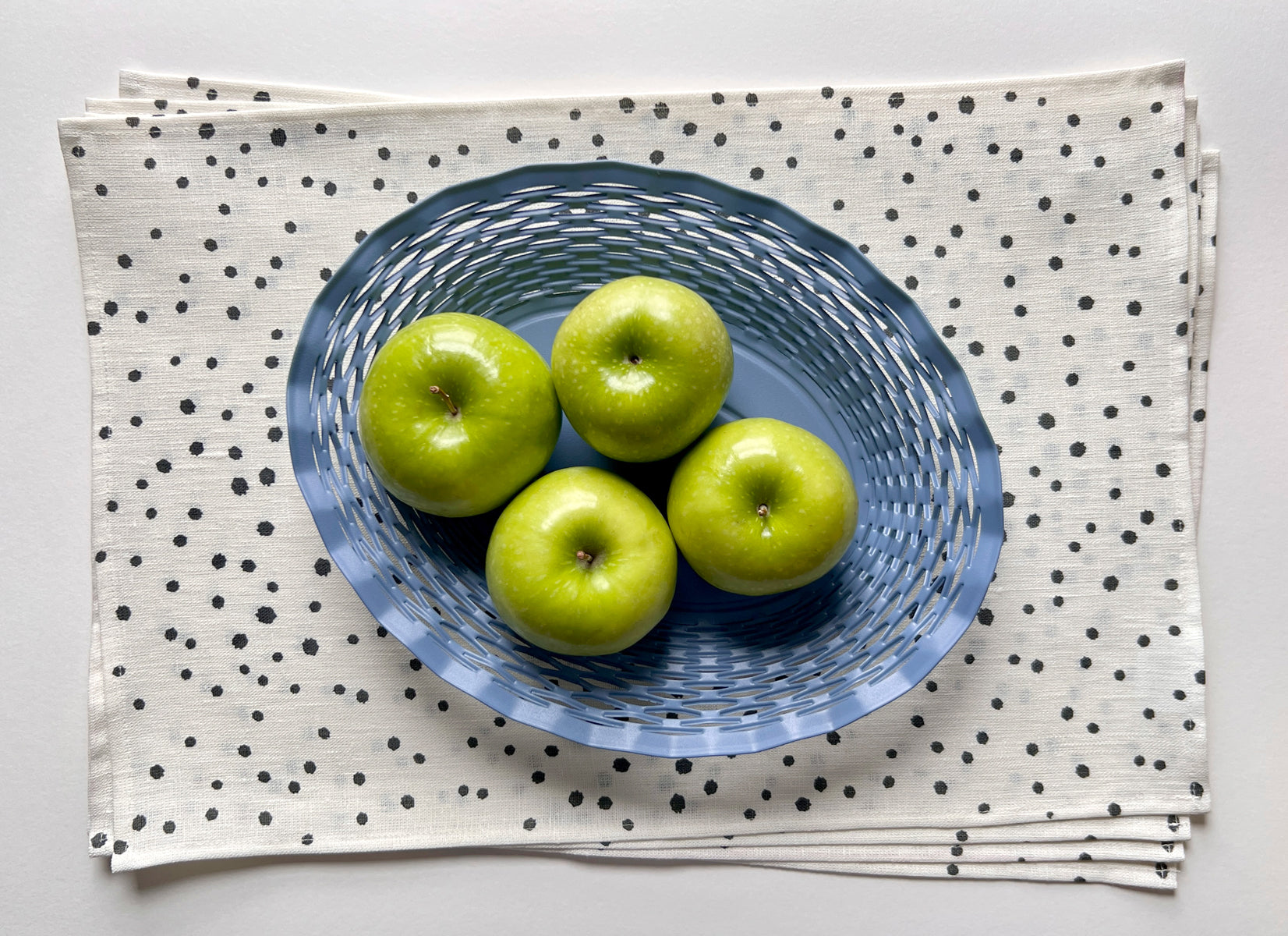 A white linen placemat with a black organic dotted pattern, displayed with a blue bowl of green apples on top. Table linens design by artist Leila Simon Hayes