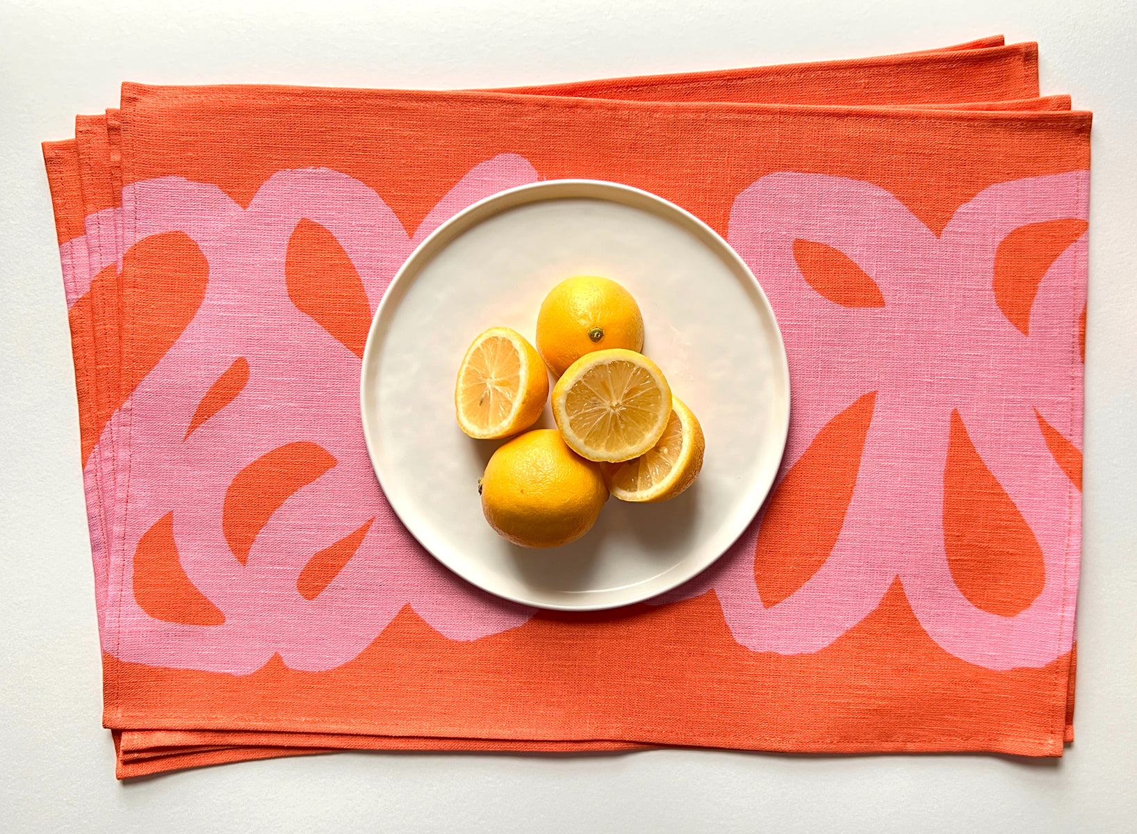 A coral colored linen placemat with an abstract pink pattern, displayed with a white plate of cut lemons on top. Designed by artist Leila Simon Hayes