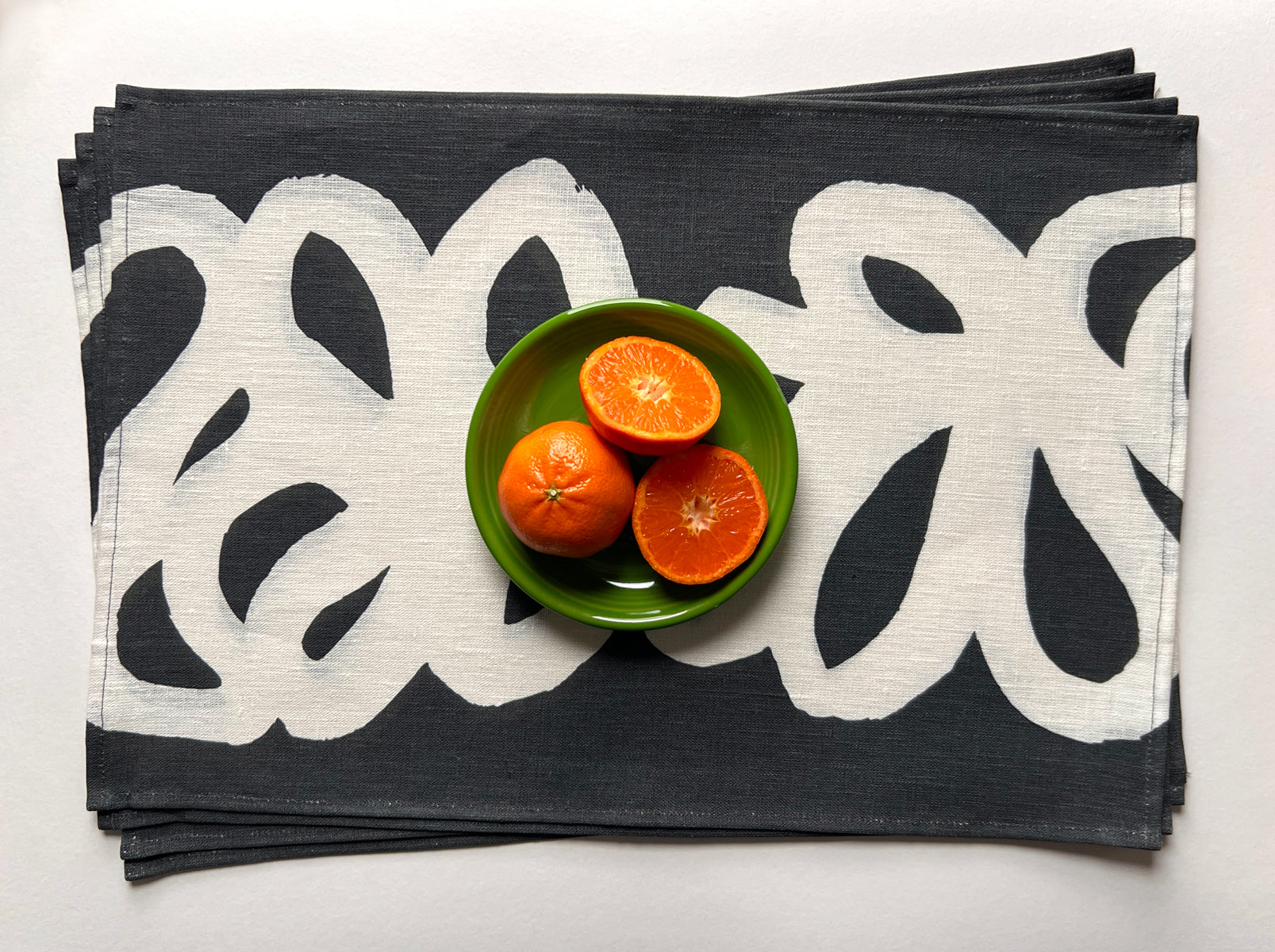 Stack of black and white patterned placemats with a green bowl containing oranges on a white background designed by artist Leila Simon Hayes