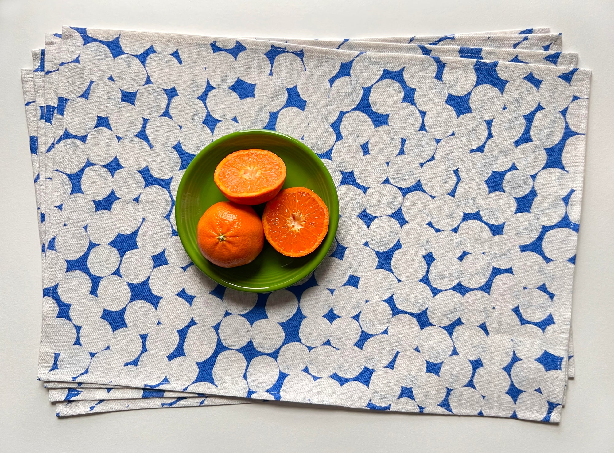 A stack of linen placemat with a sky blue color and a white dotted pattern, designed by artist Leila Simon Hayes. Displayed on a flat surface with a bowl of sliced oranges on top of it.