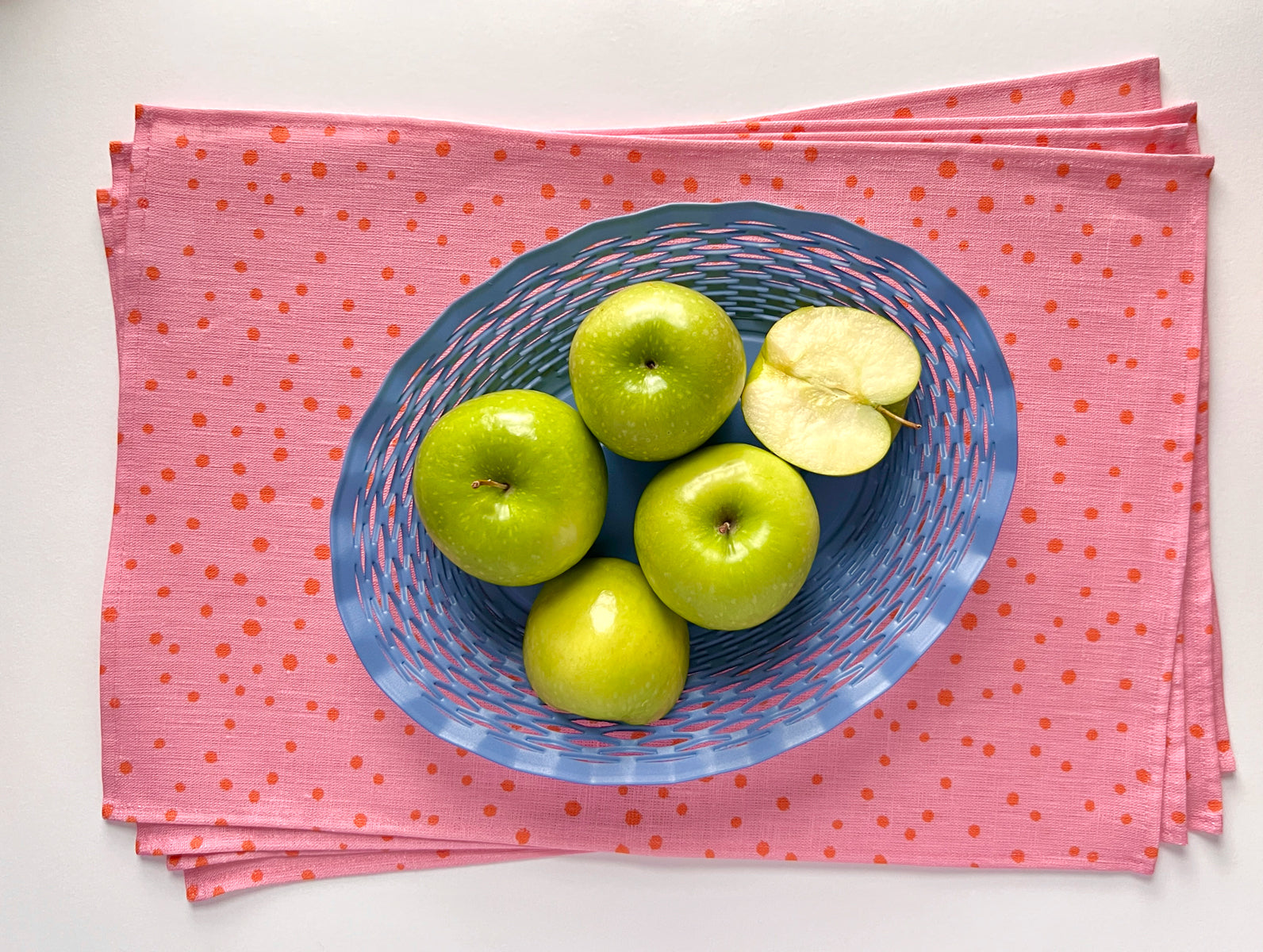 A pink linen placemat with an organic dotted pattern, displayed with a blue bowl of green apples on top. Designed by artist Leila Simon Hayes