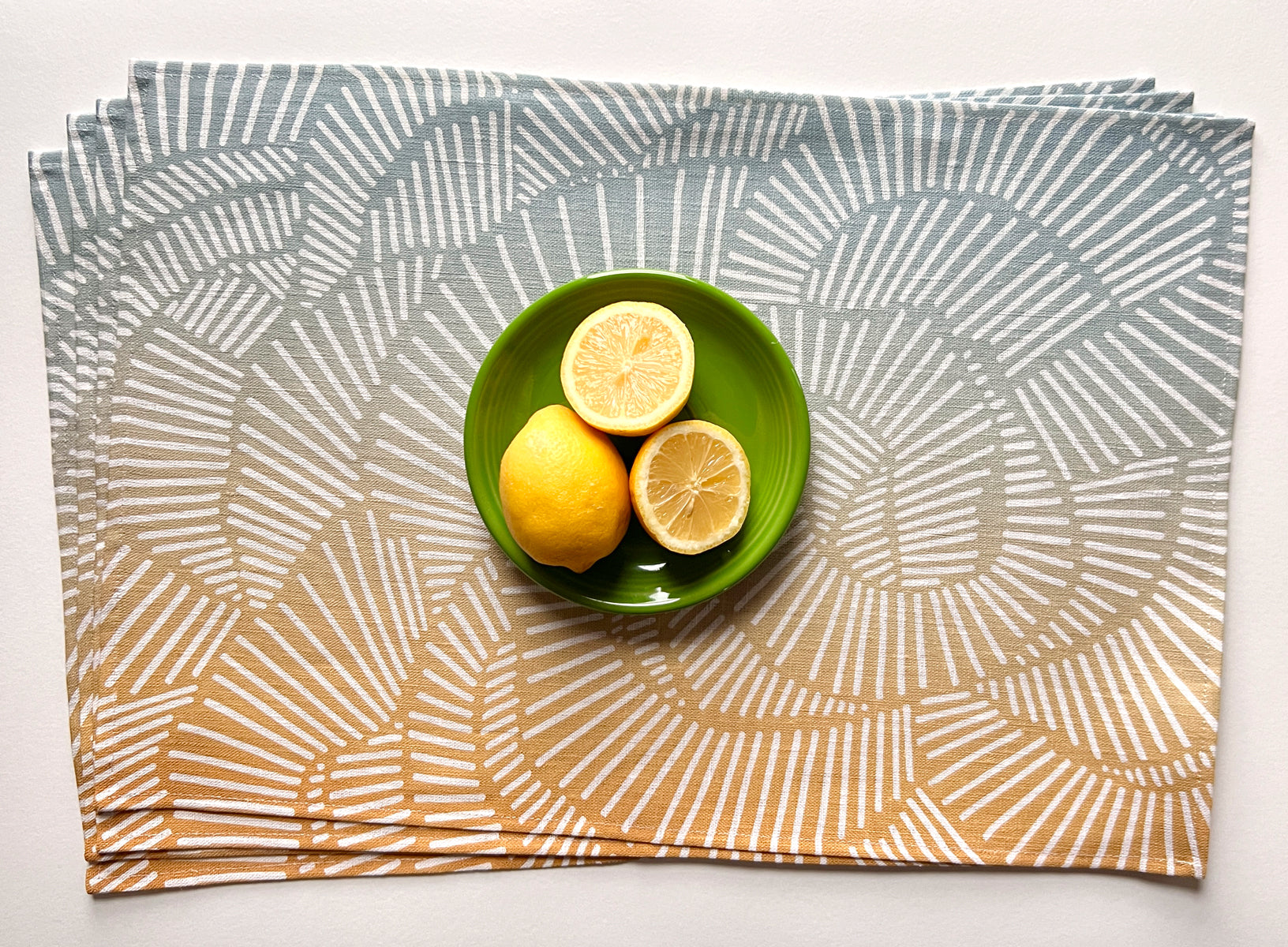 A sky blue linen placemat with an organic line pattern creating abstract shapes, displayed under natural light, with a small green bowl containing two lemon halves on top of it. Designed by artist Leila Simon Hayes