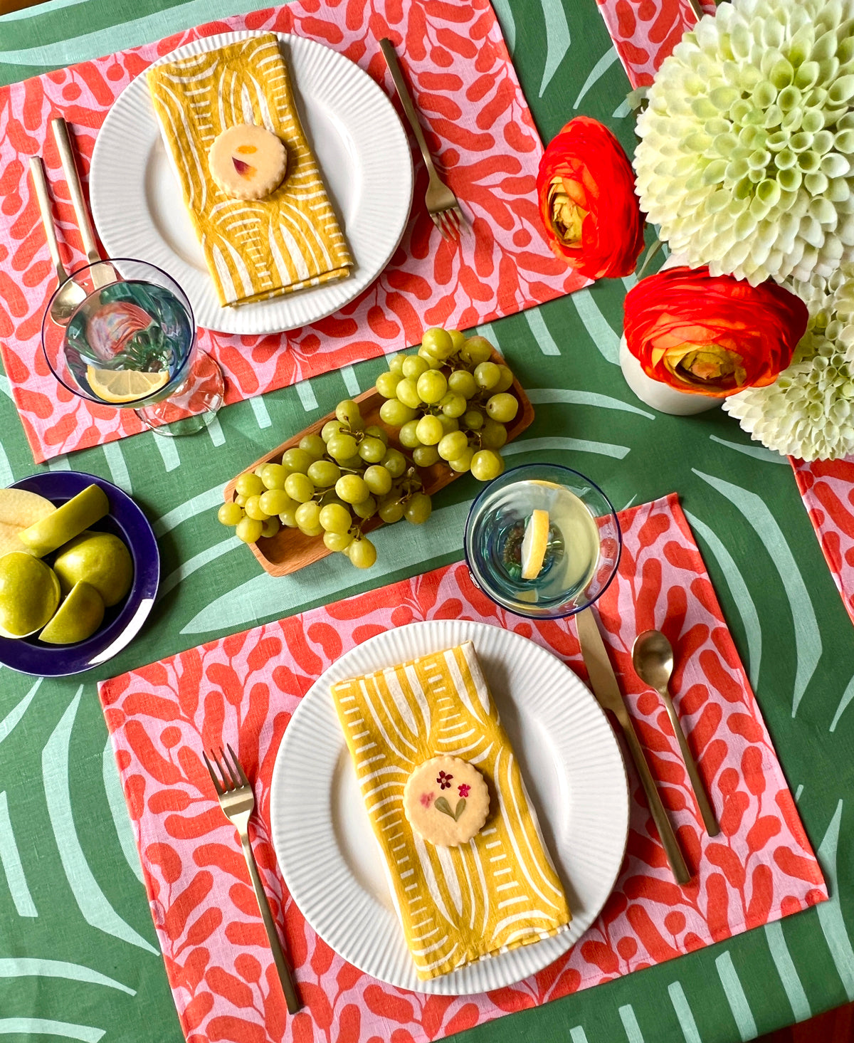 Colorful table setting with plates, yellow striped napkins, pink and coral floral placemats and a green geometrical striped tablecloth. Designed by artist Leila Simon Hayes