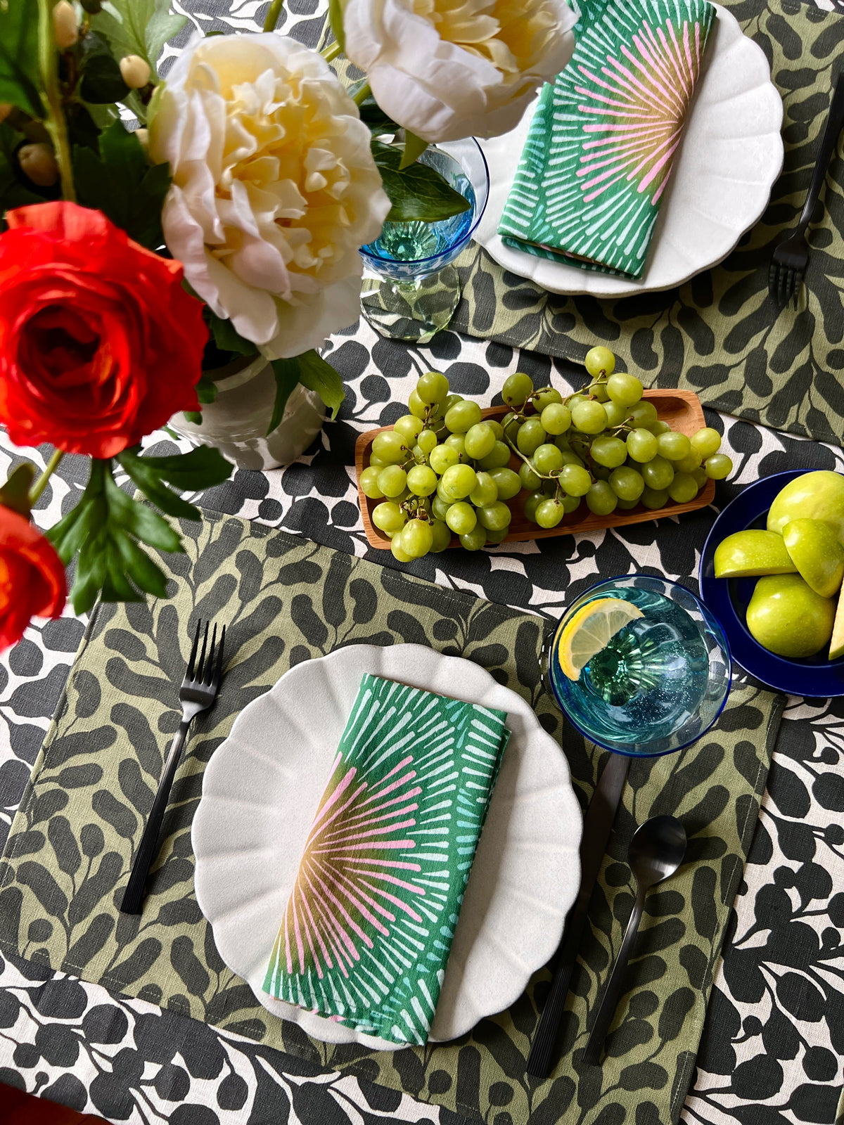 Decorative table setting with flowers, grapes, and colorful green napkins on a olive green botanical leaf patterned placemat, with a black and white floral tablecloth. Designed by artist Leila Simon Hayes