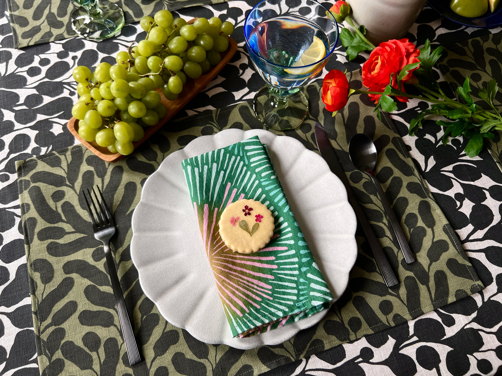 Decorative table setting with flowers, grapes, and colorful green napkins on a olive green botanical leaf patterned placemat, with a black and white floral tablecloth. Designed by artist Leila Simon Hayes