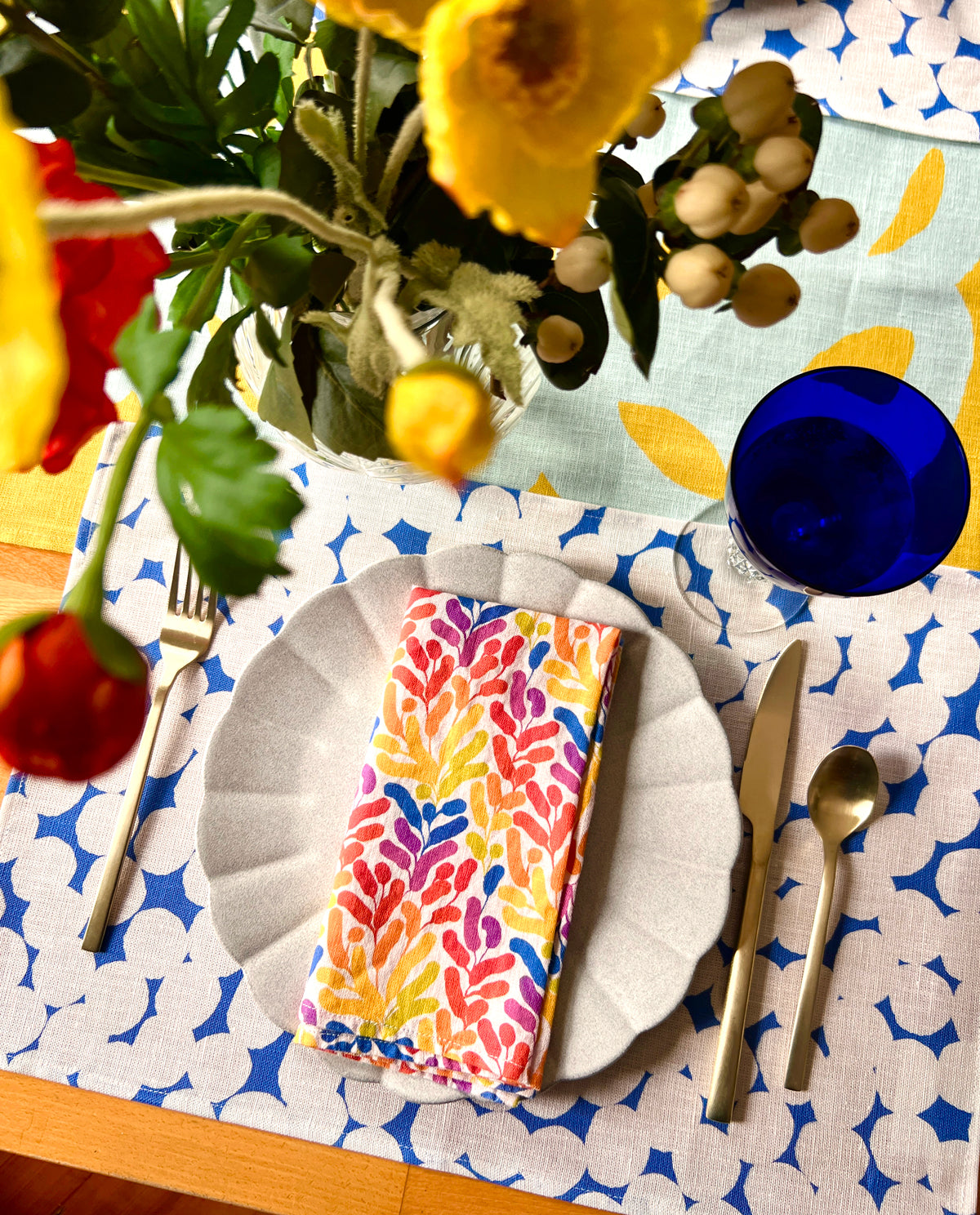 Colorful table setting with a floral centerpiece, rainbow napkin, and blue and white dots patterned placemat. Designed by artist Leila Simon Hayes