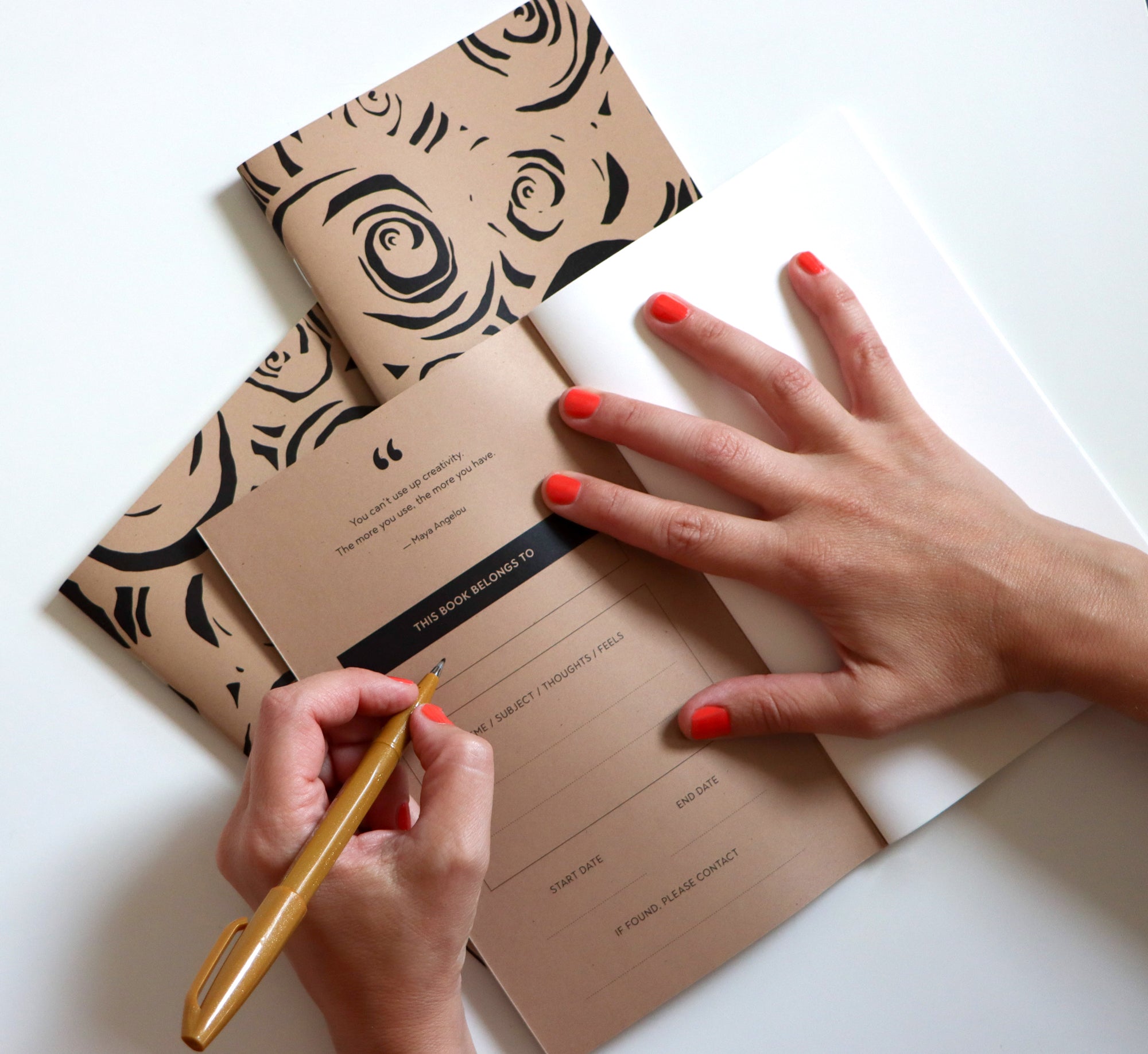 Person holding a pen over a brown notebook with rose pattern on a white surface. Designed by artist Leila Simon Hayes.