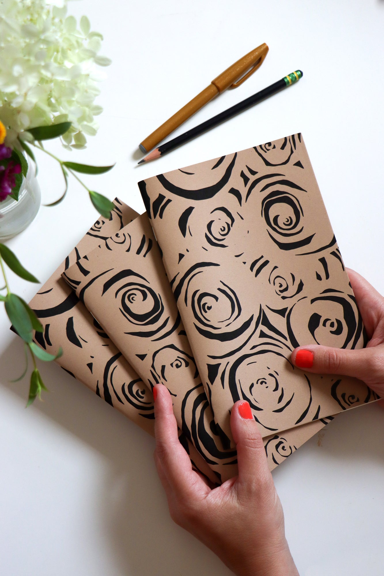 Two notebooks with black rose patterns held by a hand on a white surface with flowers and pencils. Designed by artist Leila Simon Hayes.