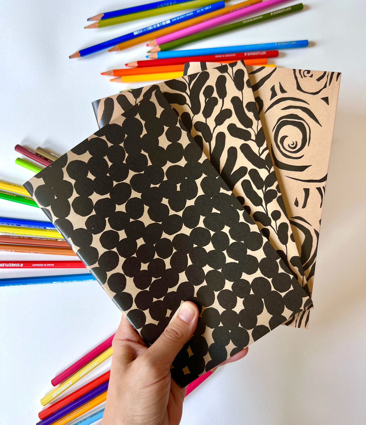 Three patterned notebooks held by a hand with colored pencils scattered around on a white background. Designed by artist Leila Simon Hayes.
