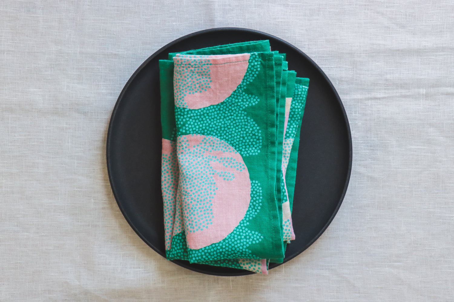 Four green folded linen napkin with a floral pattern displayed on a black plate over a white tablecloth. Designed by artist Leila Simon Hayes. 