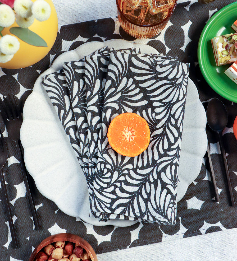 A folded black and white patterned cloth napkin with a citrus slice in the center, arranged on a white plate over a black-and-white patterned tablecloth in a styled dining setting. Designed by artist Leila Simon Hayes. 