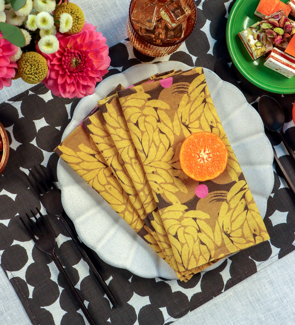 A stack of mustard yellow paper napkins with a brown leaf pattern, arranged on a white plate with a slice of orange and pink garnish, set on a black and white patterned placemat with dining utensils and flowers. Designed by artist Leila Simon Hayes. 