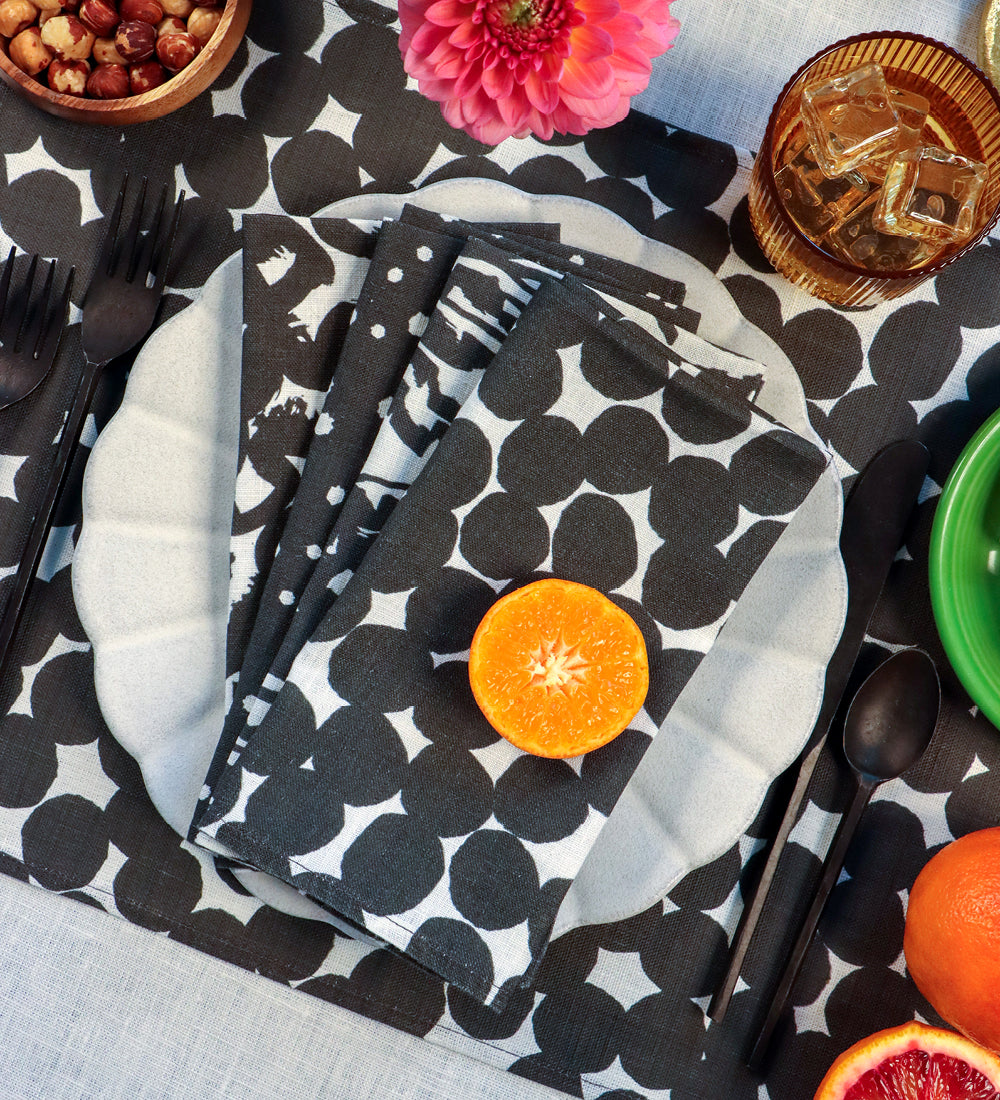 A styled tabletop setting featuring black and white polka-dot napkins on a matching placemat, with white scalloped plates, black cutlery, citrus slices, a pink flower, and a glass of iced tea. Designed by artist Leila Simon Hayes. 