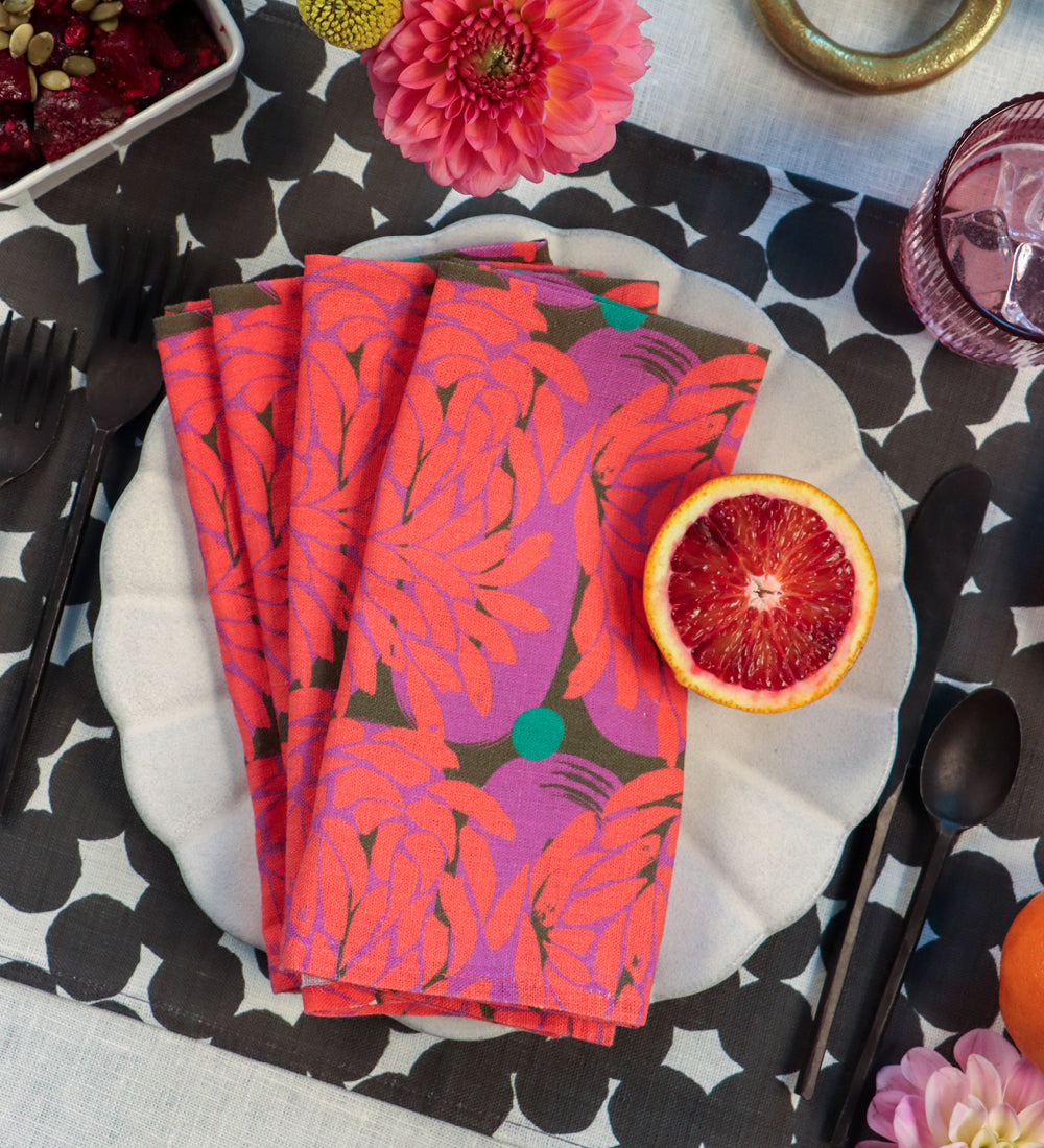 A stack of folded patterned napkins in bright pink, purple, and orange tones with small green accents, arranged on a white plate at a styled table setting with cutlery, flowers, and a sliced blood orange. Designed by artist Leila Simon Hayes. 
