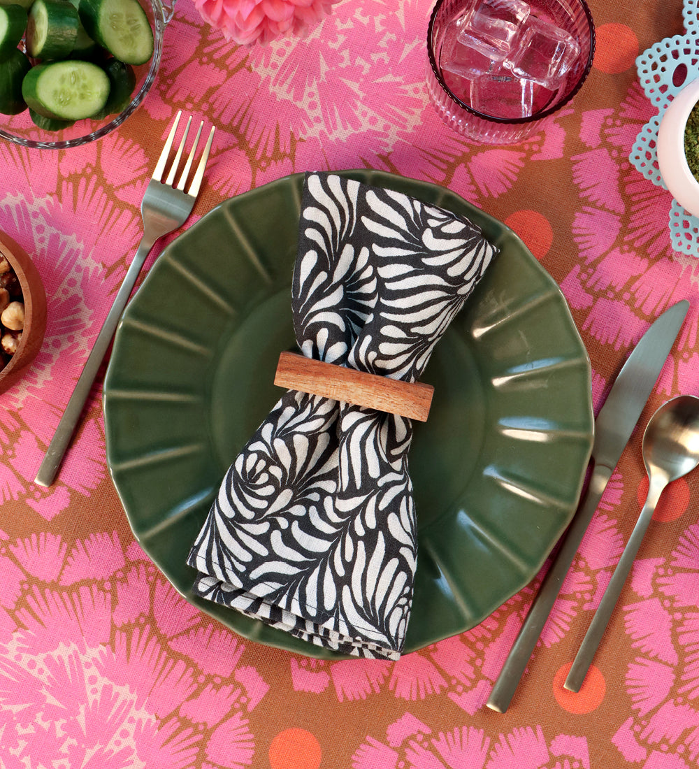 A styled table setting featuring a green ceramic plate with a folded black and white patterned linen napkin secured by a wooden ring, surrounded by silver cutlery on a pink and orange patterned tablecloth. Designed by artist Leila Simon Hayes. 