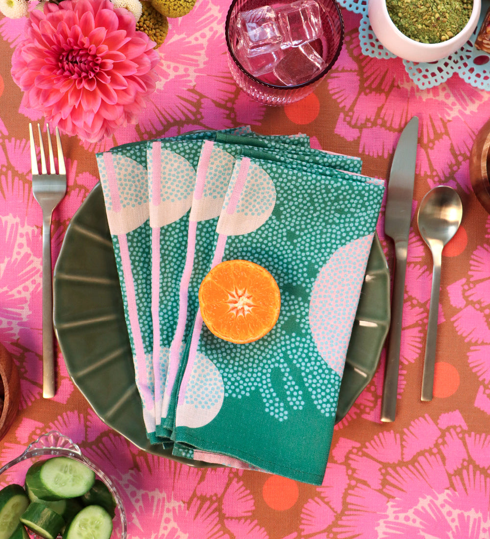 A styled tabletop setting with a green plate holding a folded teal napkin printed with white abstract shapes and pink dotted stripes. A sliced orange rests on the napkin, surrounded by silver cutlery, a pink patterned tablecloth, flowers, a glass of water, and small bowls of food. Designed by artist Leila Simon Hayes. 