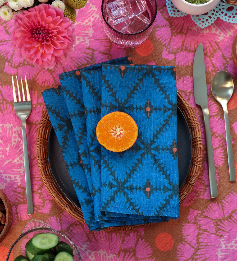 A set of folded blue patterned napkins with a dark geometric design, styled on a dark plate with a sliced orange on top, set on a pink patterned tablecloth with silver cutlery. Designed by artist Leila Simon Hayes. 