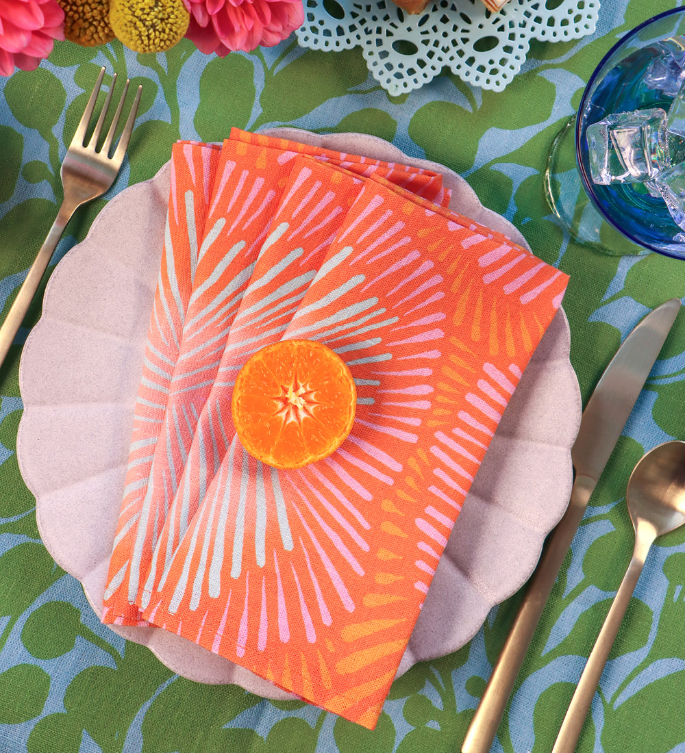 Designed by artist Leila Simon Hayes. Orange sunburst napkins on a white plate with a botanical green and blue tablecloth.