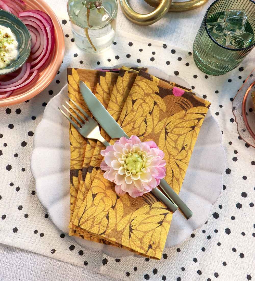 A styled tabletop setting featuring a folded napkin with a mustard yellow and brown botanical leaf pattern, set on a white scalloped plate with silver cutlery and a pink flower accent. Designed by artist Leila Simon Hayes. 