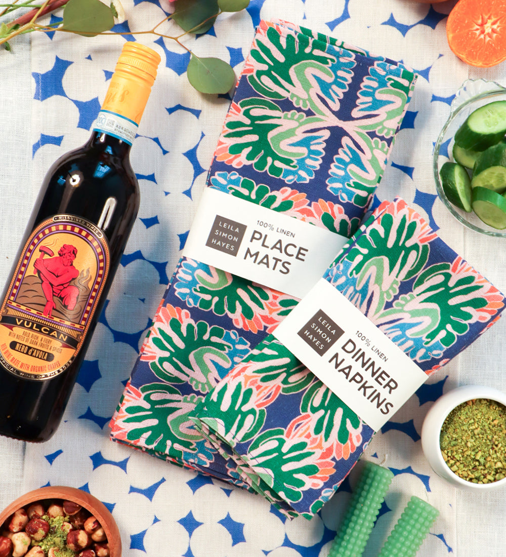 Two patterned table linens from Leila Simon Hayes are arranged on a blue-and-white patterned tablecloth with a bottle of red wine nearby. The place mats and dinner napkins have a tropical leaf print in green, pink, and blue, with white paper bands showing the brand names. Designed by artist Leila Simon Hayes. 