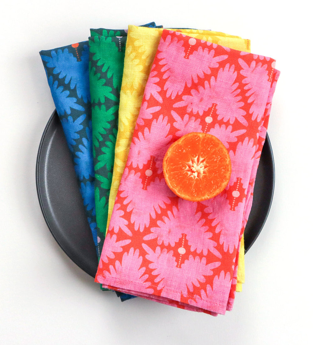 Four folded patterned cloth napkins in blue, green, yellow, and pink-red are stacked on a dark round plate, with a sliced orange placed on top. The napkins feature a repeating leaf-like floral pattern. Designed by artist Leila Simon Hayes. 