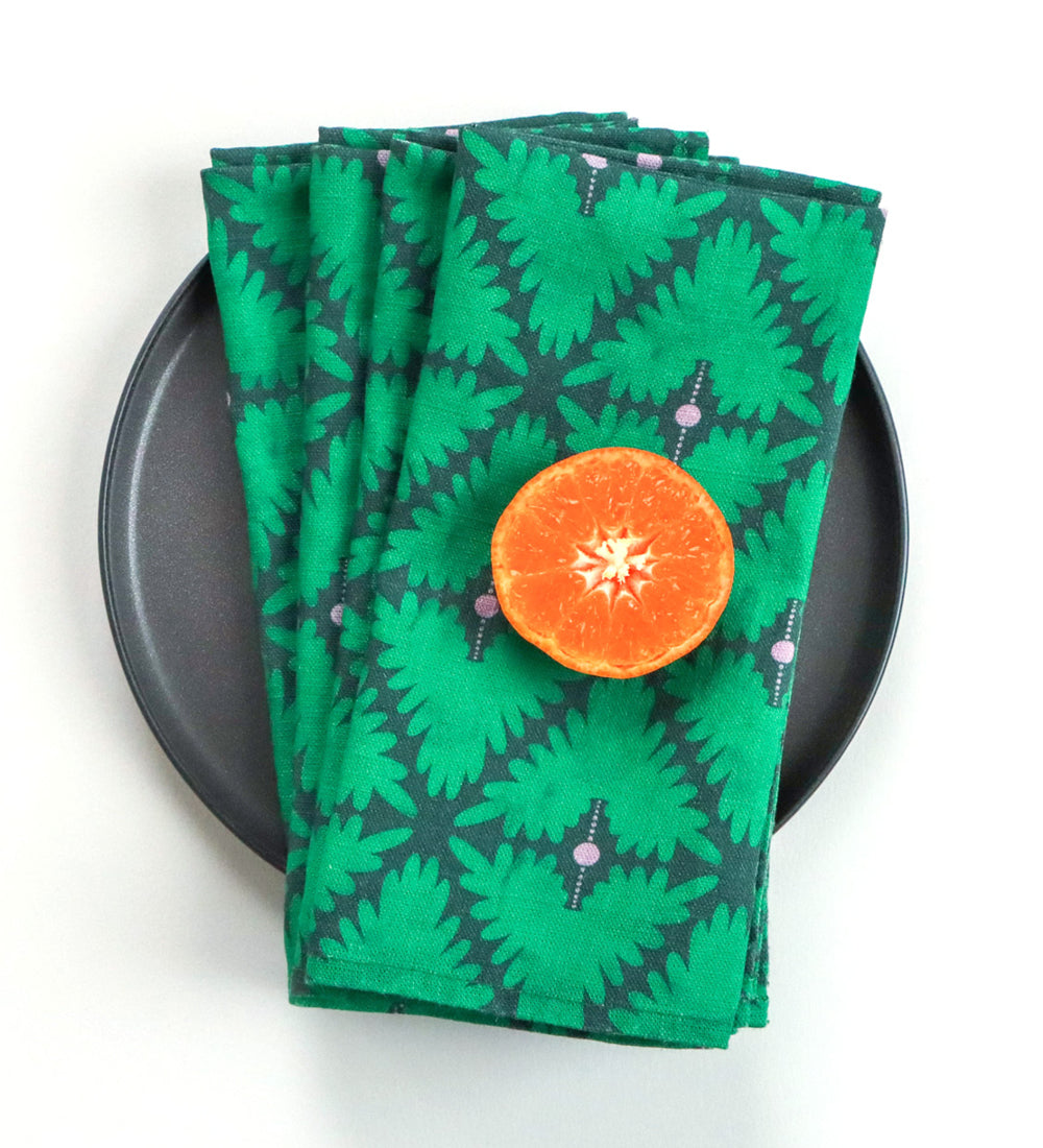 Designed by artist Leila Simon Hayes. Four linen dinner napkins in a green hand-drawn design on a black plate with an orange on top.