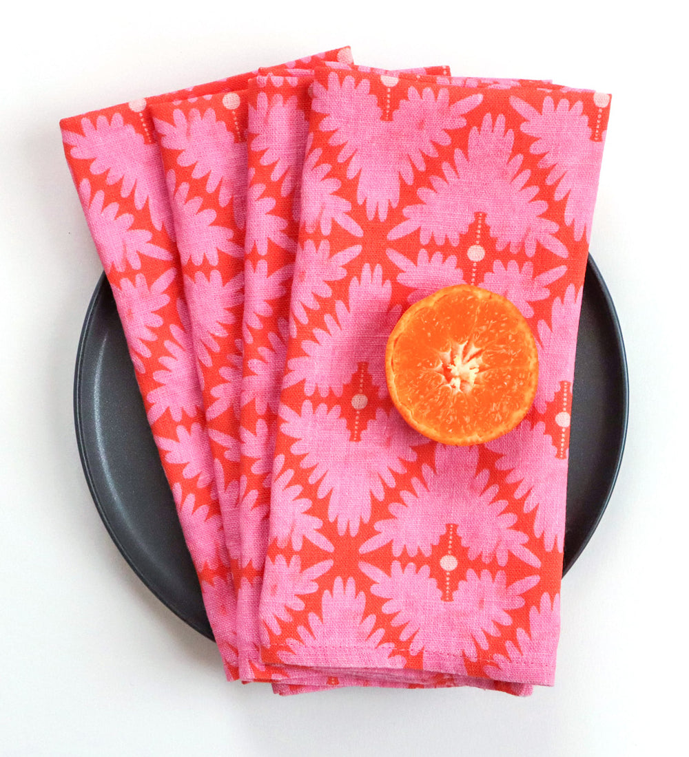 A stack of pink and orange patterned cloth napkins with a cut orange half placed on top, arranged on a round black plate against a white background. Designed by artist Leila Simon Hayes. 