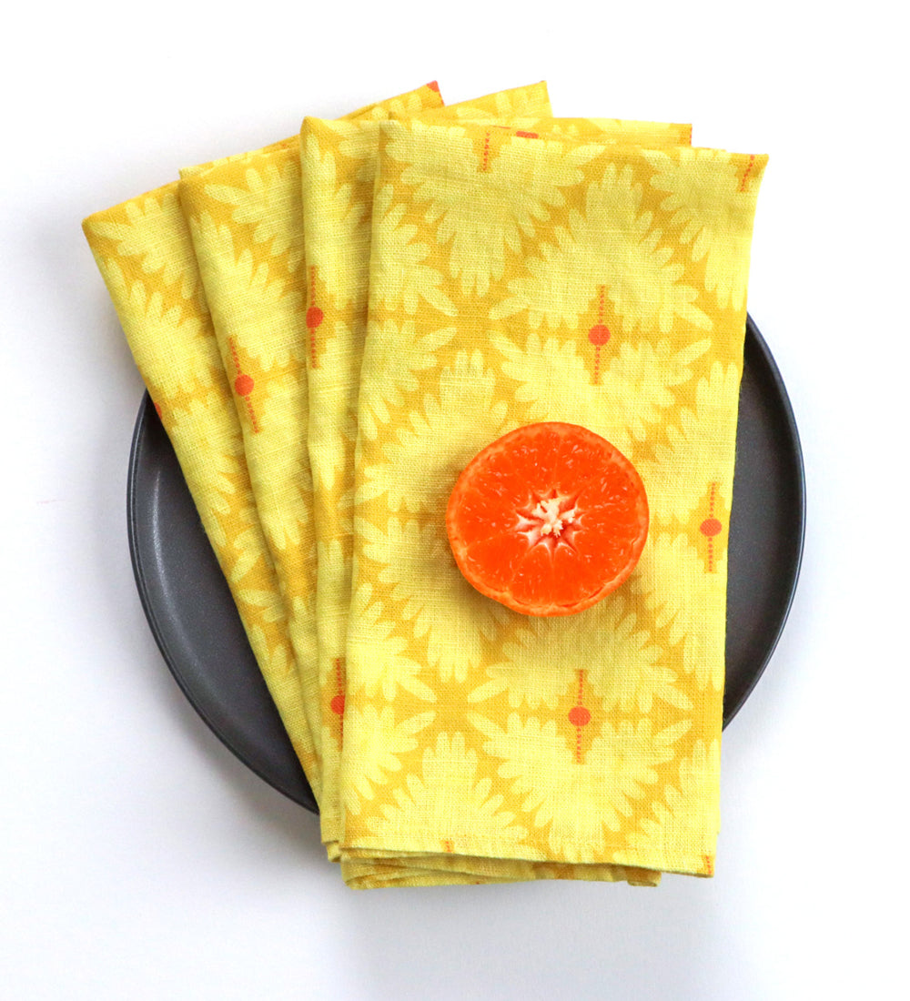 A stack of yellow patterned cloth napkins with a mandala design and small orange dots, arranged on a dark gray plate with a halved orange on top, against a white background. Designed by artist Leila Simon Hayes. 
