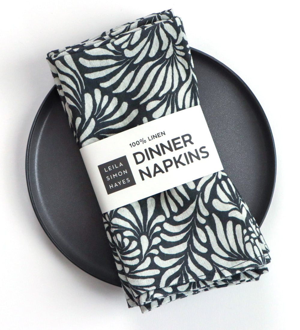 Folded linen napkins with a black and white botanical leaf pattern, wrapped in a white paper band that reads '100% LINEN DINNER NAPKINS' displayed on a matte black plate against a white background. Designed by artist Leila Simon Hayes. 