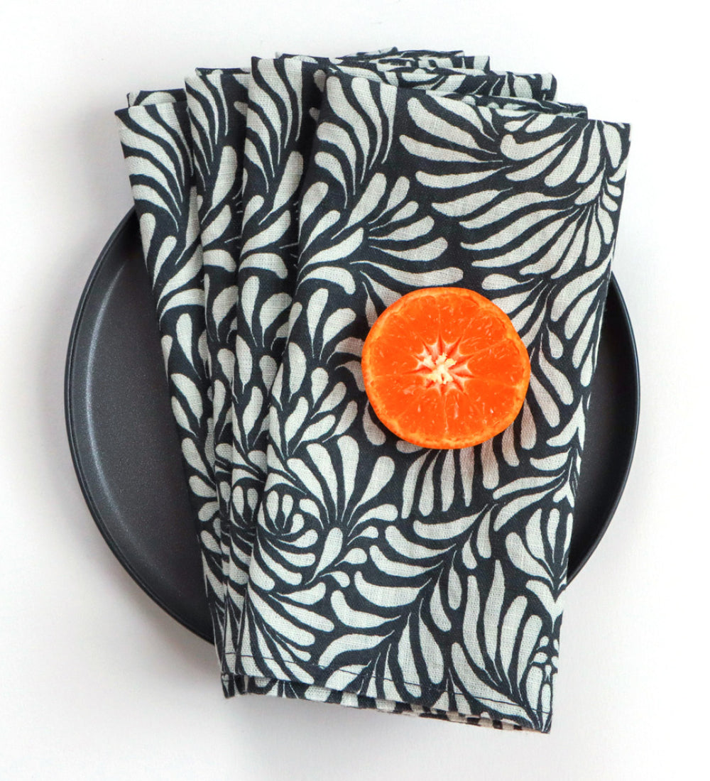 Four folded cloth napkins with a black and white floral pattern, styled on a black plate with a halved orange slice on top, against a plain white background.Designed by artist Leila Simon Hayes. 