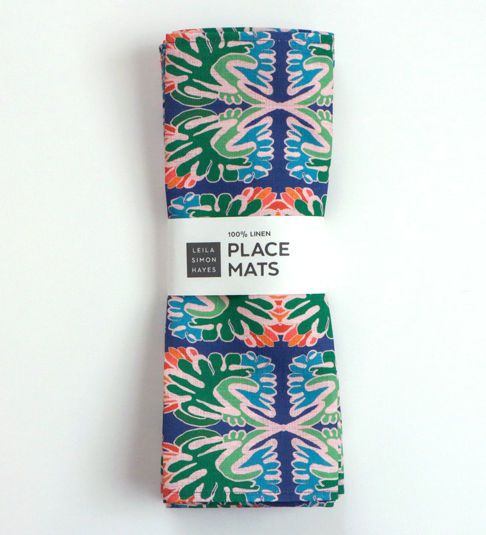 A folded set of two patterned linen place mats with a colorful mandala design on a navy blue background, wrapped in a white paper band. Designed by artist Leila Simon Hayes. 