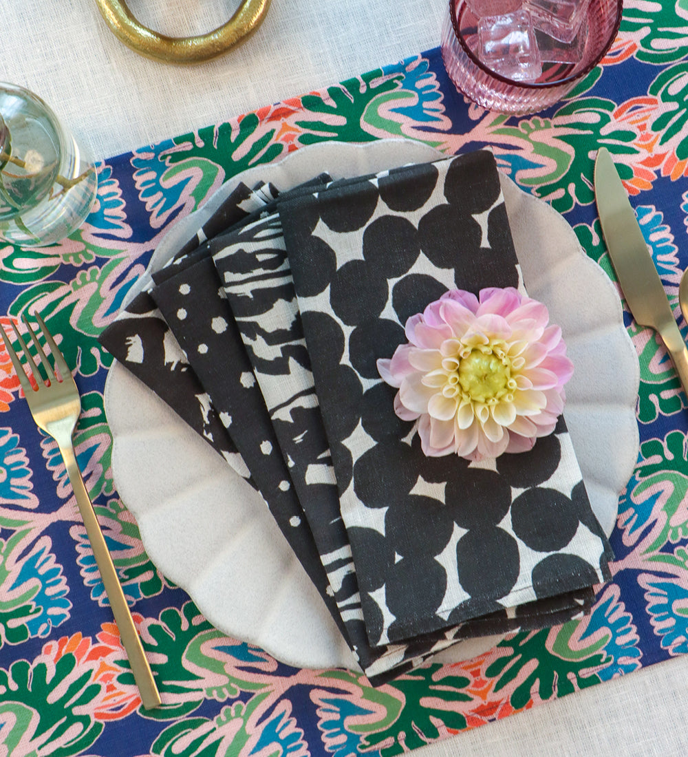 A styled tabletop setting featuring a stack of black and white patterned cloth napkins on a white plate, decorated with a pink flower. The napkins are placed on a colorful floral placemat with gold-toned cutlery and glassware around them. Designed by artist Leila Simon Hayes. 