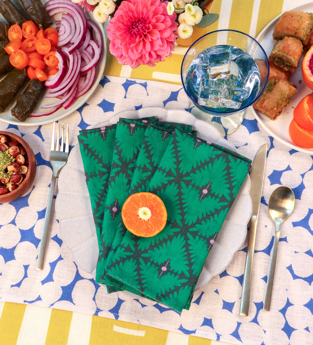 A styled table setting featuring a stack of green patterned paper napkins with a central orange slice garnish, surrounded by plates of food, silverware, and a blue-and-white patterned tablecloth. Designed by artist Leila Simon Hayes. 