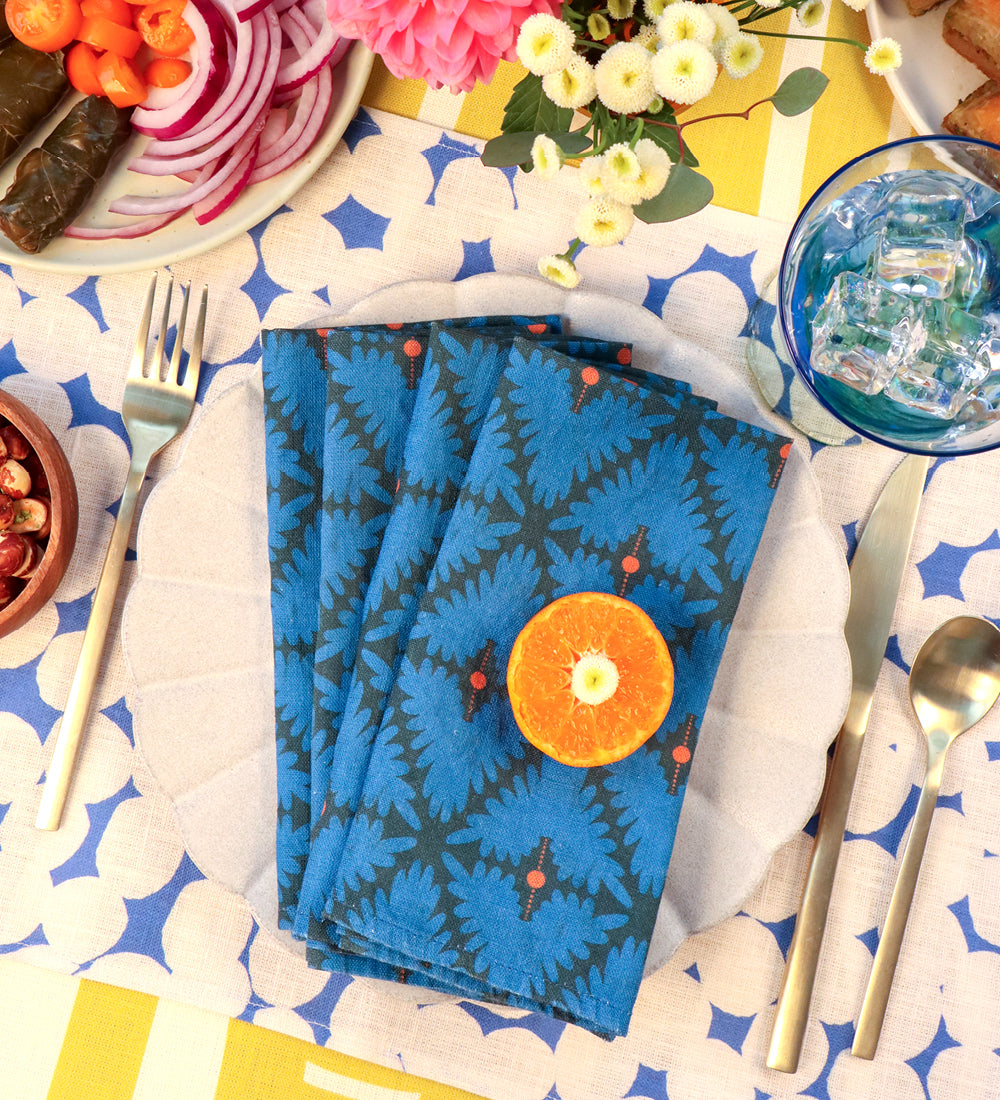 A stack of blue patterned cloth napkins with a dark fern-like print and small red berry accents, styled on a white plate at a table setting. An orange slice rests on top, with silverware, a blue drink, flowers, sliced red onion, and other food visible around it. Designed by artist Leila Simon Hayes. 