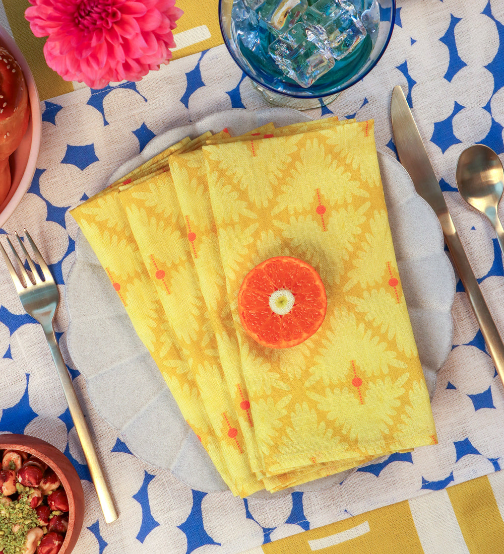 A stack of yellow patterned cloth napkins with orange accents, arranged on a white plate at a table setting. A slice of orange sits on top, with a fork, knife, spoon, blue drink, flowers, and food visible around it. Designed by artist Leila Simon Hayes. 