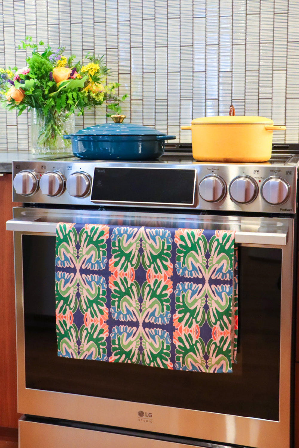 An over with a colorful patterned kitchen towel hanging on the oven door handle. A blue Dutch oven and a yellow pot sit on the stovetop, with a tiled backsplash and a vase of flowers in the background. Designed by artist Leila Simon Hayes.
