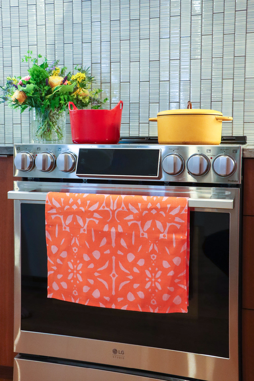 An over with a colorful patterned kitchen towel hanging on the oven door handle. A blue Dutch oven and a yellow pot sit on the stovetop, with a tiled backsplash and a vase of flowers in the background. Designed by artist Leila Simon Hayes.
