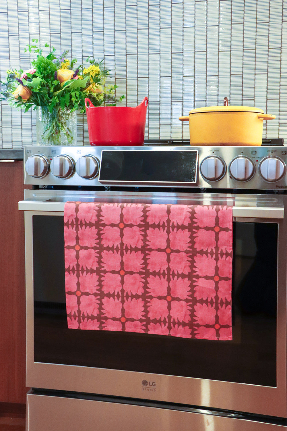 An over with a colorful patterned kitchen towel hanging on the oven door handle. A blue Dutch oven and a yellow pot sit on the stovetop, with a tiled backsplash and a vase of flowers in the background. Designed by artist Leila Simon Hayes.