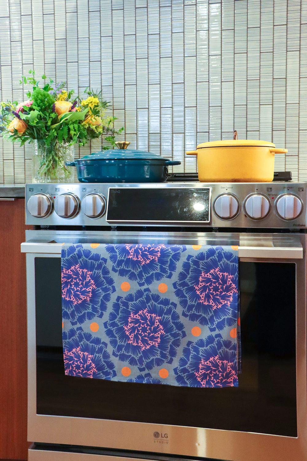 An over with a colorful patterned kitchen towel hanging on the oven door handle. A blue Dutch oven and a yellow pot sit on the stovetop, with a tiled backsplash and a vase of flowers in the background. Designed by artist Leila Simon Hayes.