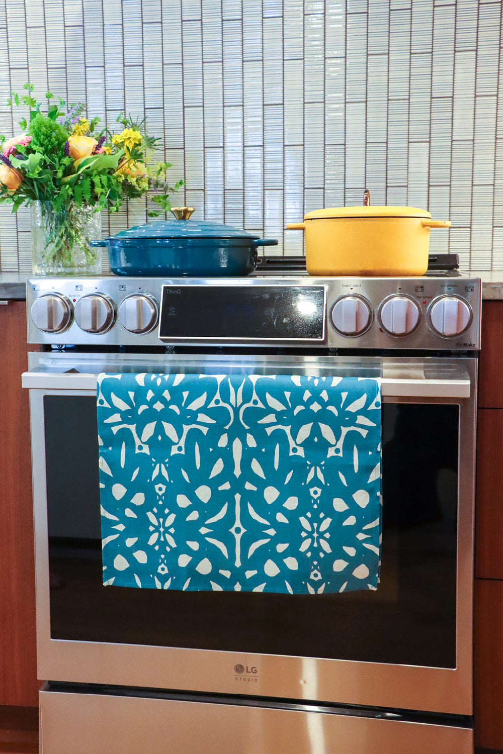 An over with a colorful patterned kitchen towel hanging on the oven door handle. A blue Dutch oven and a yellow pot sit on the stovetop, with a tiled backsplash and a vase of flowers in the background. Designed by artist Leila Simon Hayes.