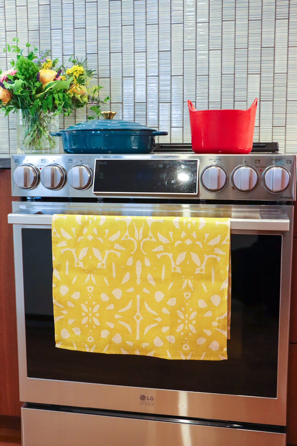 An over with a colorful patterned kitchen towel hanging on the oven door handle. A blue Dutch oven and a yellow pot sit on the stovetop, with a tiled backsplash and a vase of flowers in the background. Designed by artist Leila Simon Hayes.