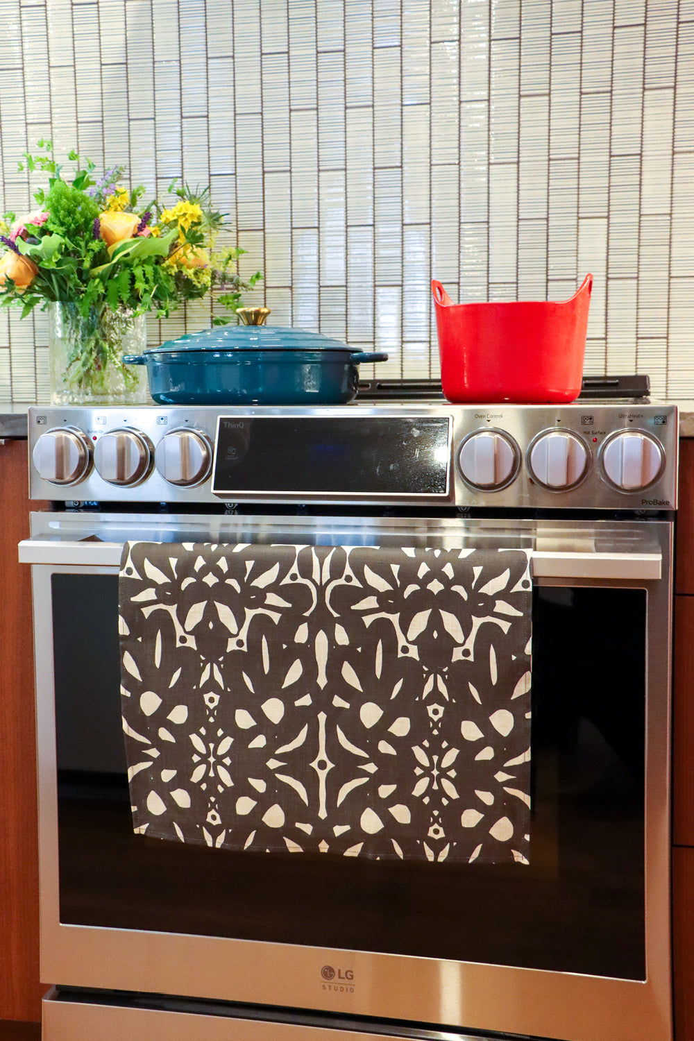 An over with a colorful patterned kitchen towel hanging on the oven door handle. A blue Dutch oven and a yellow pot sit on the stovetop, with a tiled backsplash and a vase of flowers in the background. Designed by artist Leila Simon Hayes.