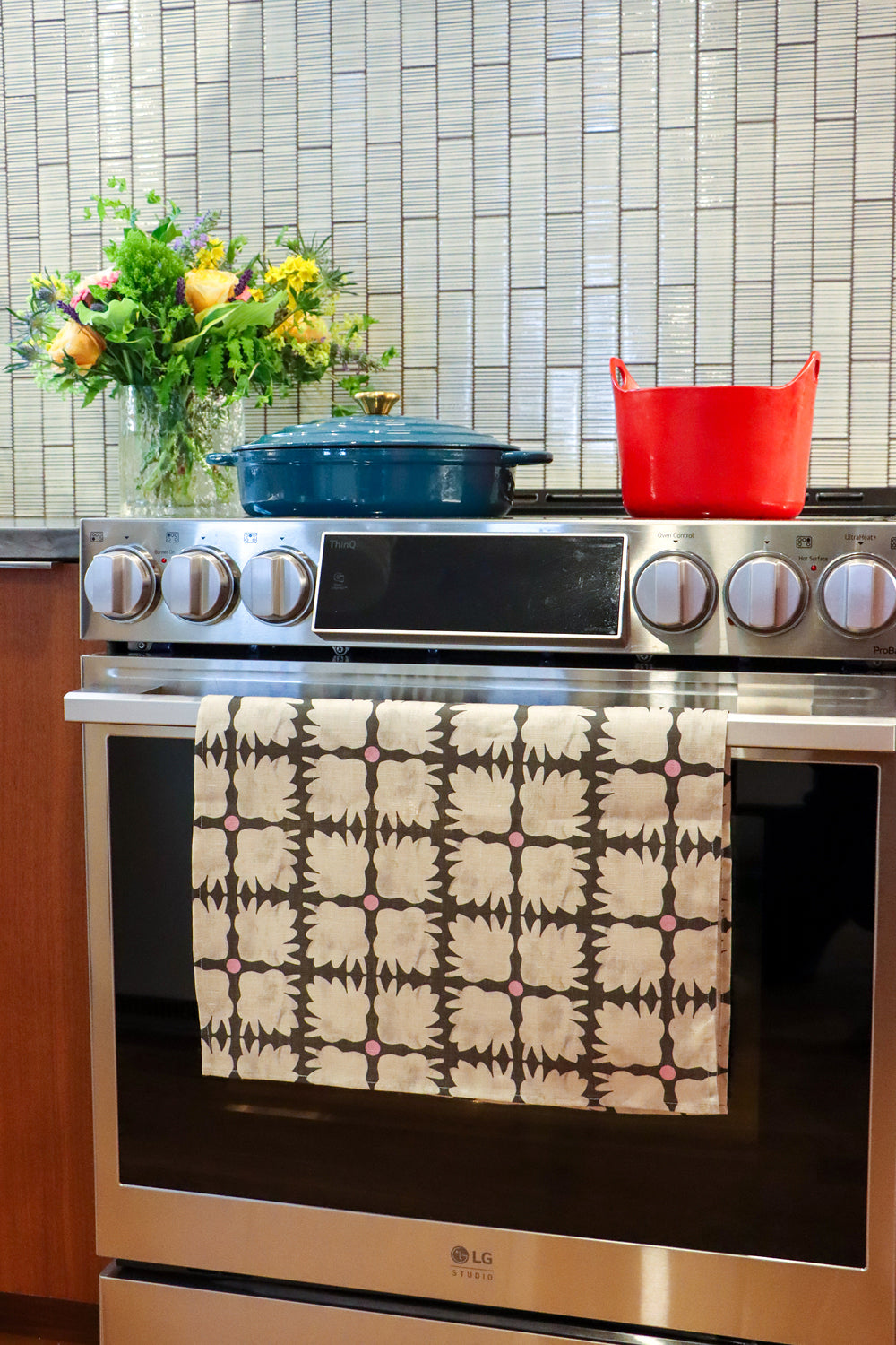 An over with a colorful patterned kitchen towel hanging on the oven door handle. A blue Dutch oven and a yellow pot sit on the stovetop, with a tiled backsplash and a vase of flowers in the background. Designed by artist Leila Simon Hayes.