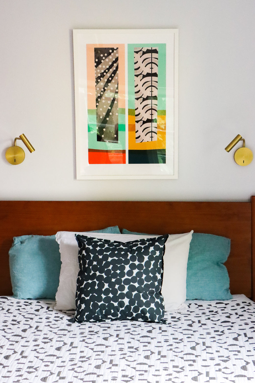 A styled bedroom scene featuring a framed abstract art print with vertical panels in coral, black, white, teal, and yellow, hung on a light gray wall above a wooden headboard with patterned pillows and a matching patterned duvet.