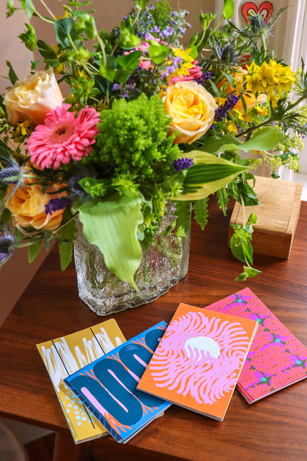 A vibrant floral arrangement in a clear glass vase on a wooden table, alongside four colorful patterned postcard packs of 10. Designed by artist Leila Simon Hayes.