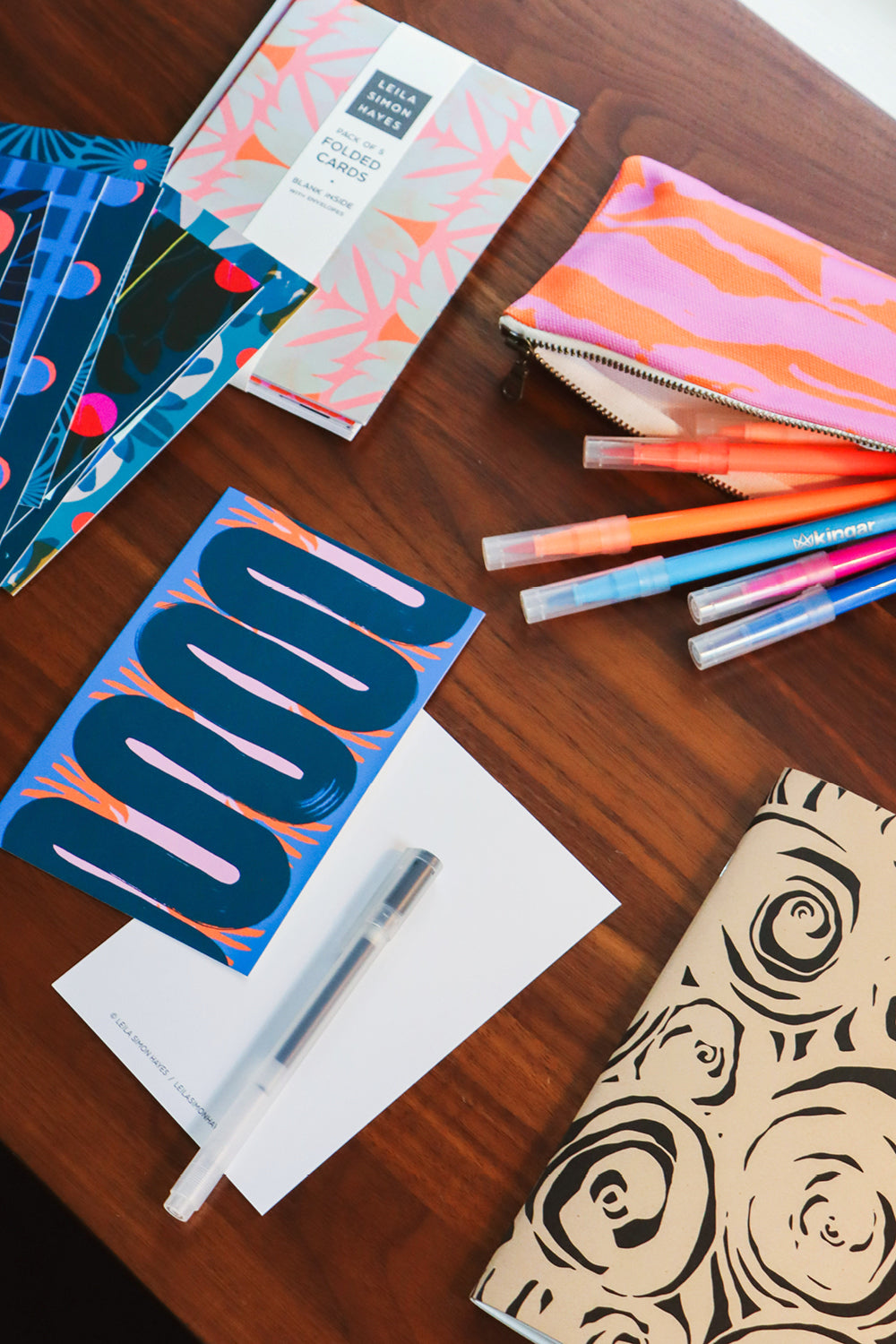 A styled flat-lay of stationery items on a wooden table, including patterned notebooks, a zip pouch, pens, and a white pen on a sheet of paper. Designed by artist Leila Simon Hayes.