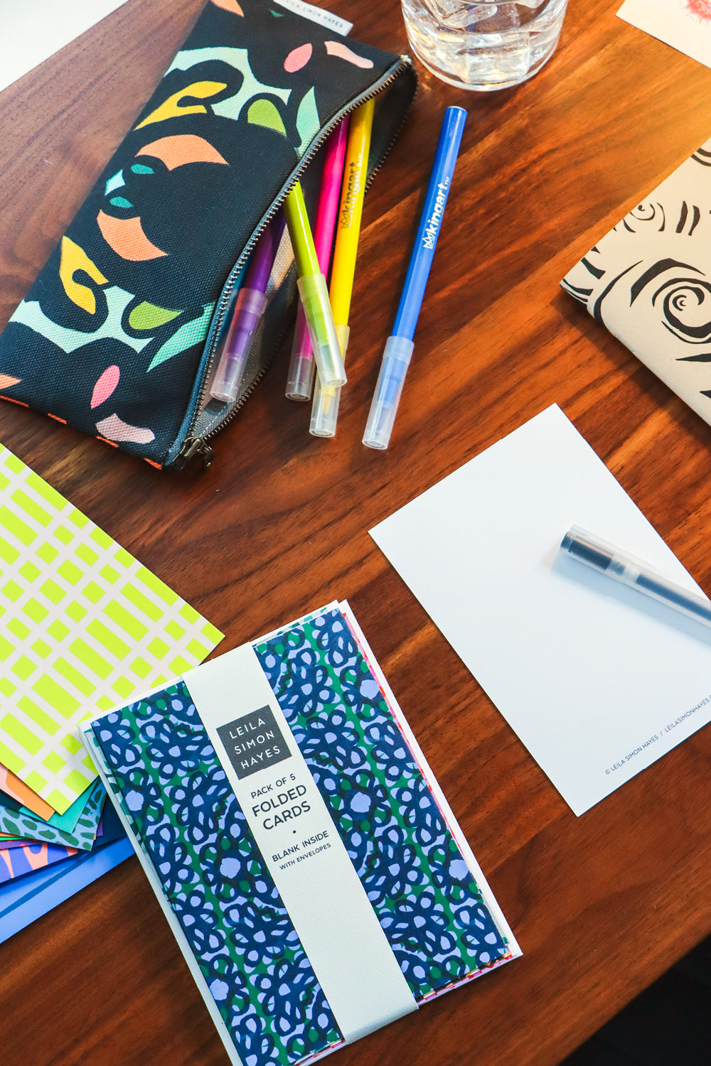 A styled flat-lay on a wooden desk featuring a patterned pencil pouch, a pack of blue and green patterned folded cards, a white notepad, and pens. Designed by artist Leila Simon Hayes.